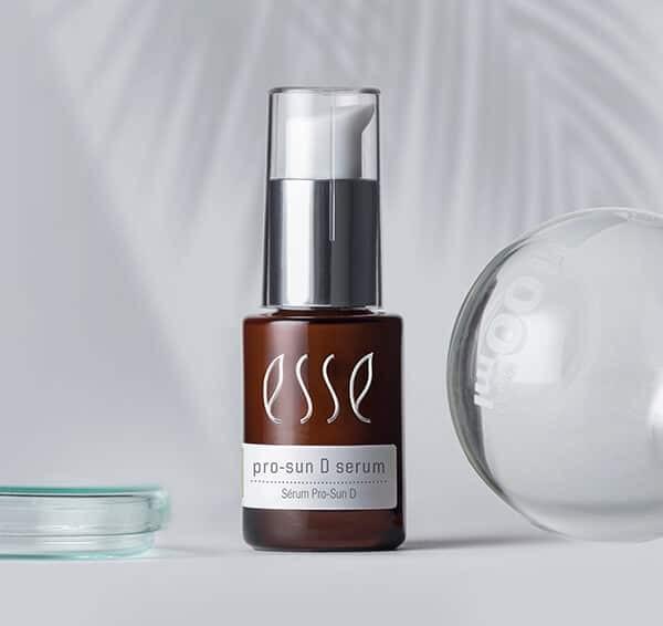 A brown bottle labeled "Esse pro-sun D serum" with a silver and white pump stands on a light surface, next to a small glass dish and a clear rounded object. Pale palm leaf shadows appear in the background.
