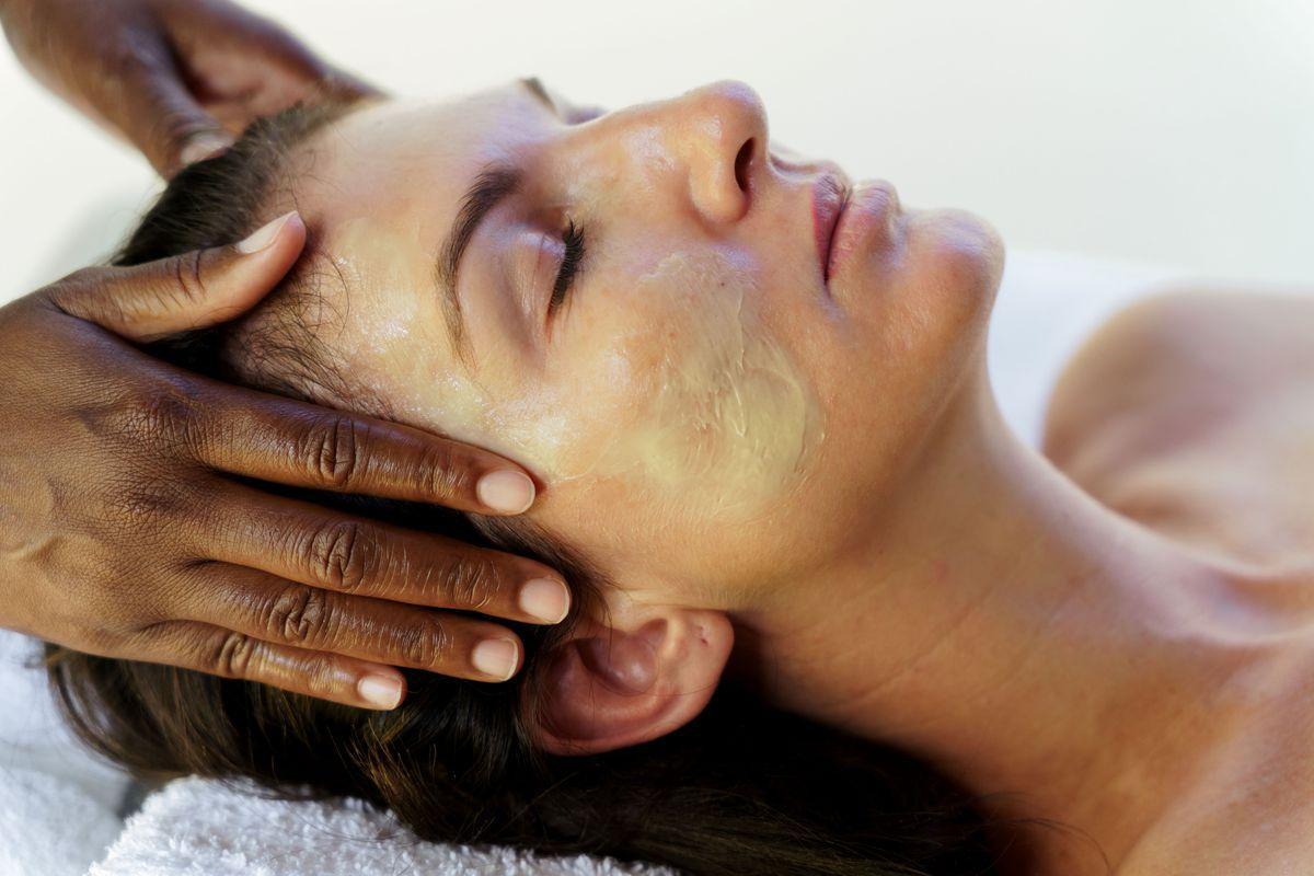 A person with a facial mask lies with eyes closed while another person massages their forehead and scalp, suggesting a relaxing skincare or spa treatment.